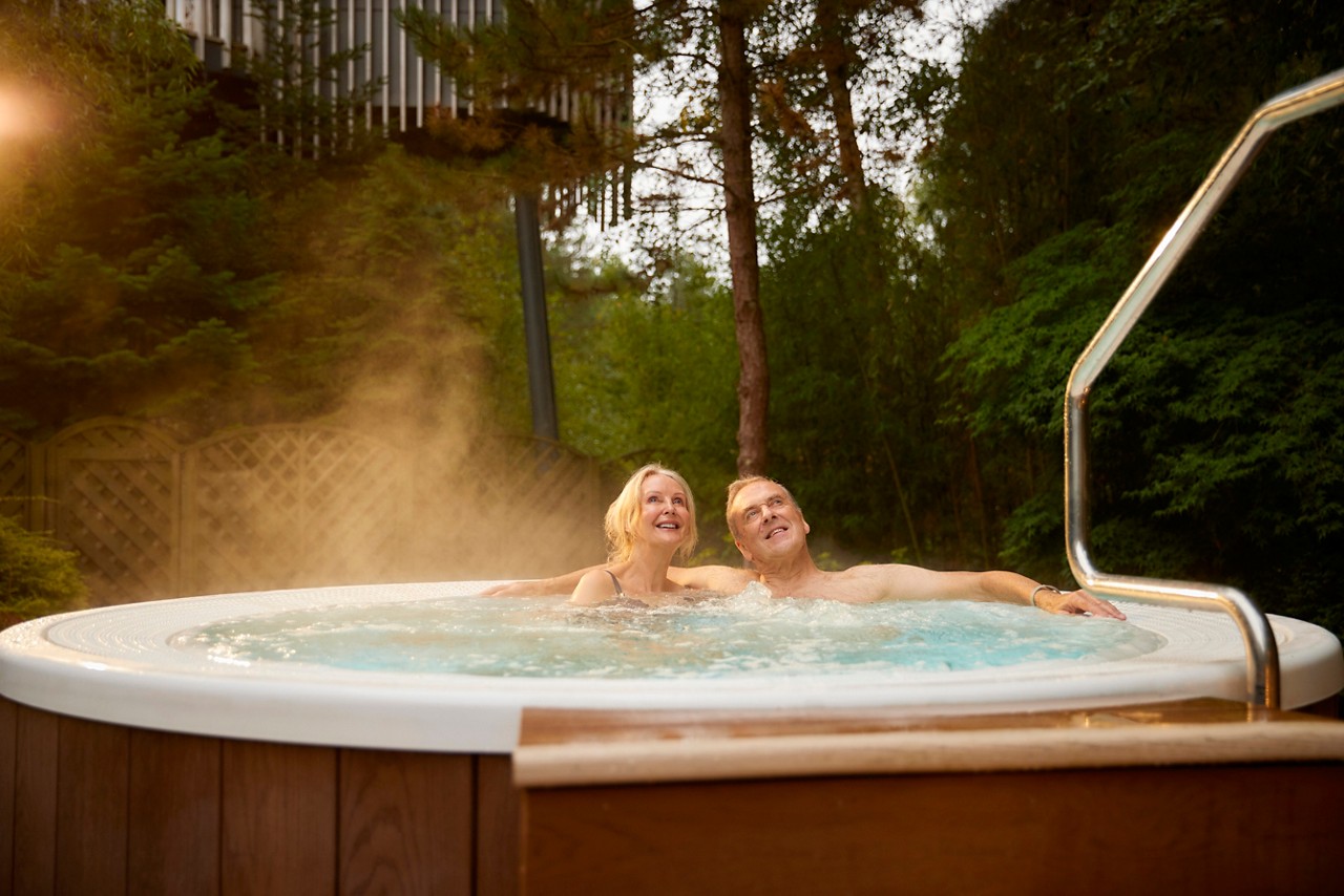 A smiling man and women relaxing in an outdoor hot tub surrounded by pine trees, looking up into the forest canopy