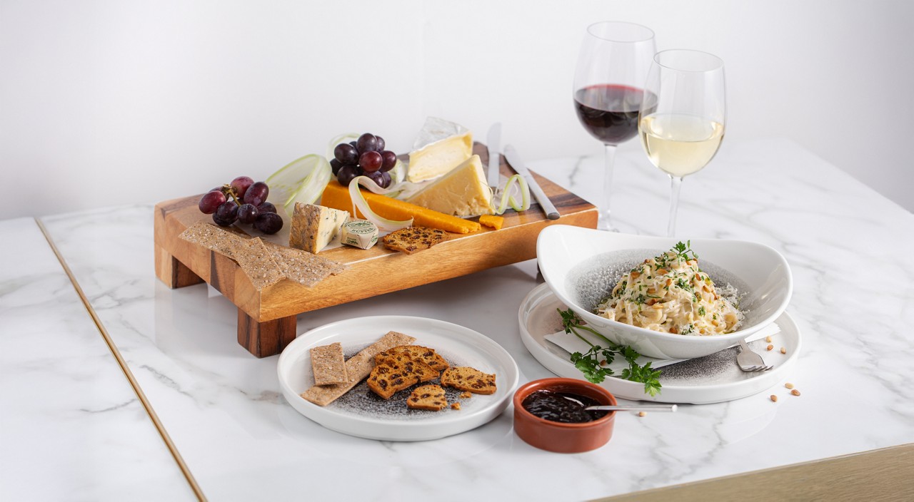 A elevated wooden cheese platter with various cheeses, grapes, and crackers, served alongside a bowl of creamy pasta, a side of jam and two glasses of wine on a marble surface