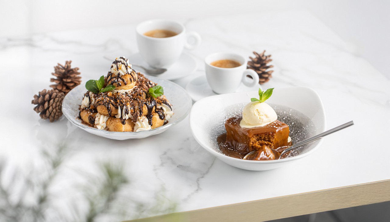 a dessert spread featuring a stack of profiteroles topped with whipped cream and chocolate sauce next to a sticky toffee pudding with vanilla ice cream served with two cups of coffee on a light table decorated with pinecones
