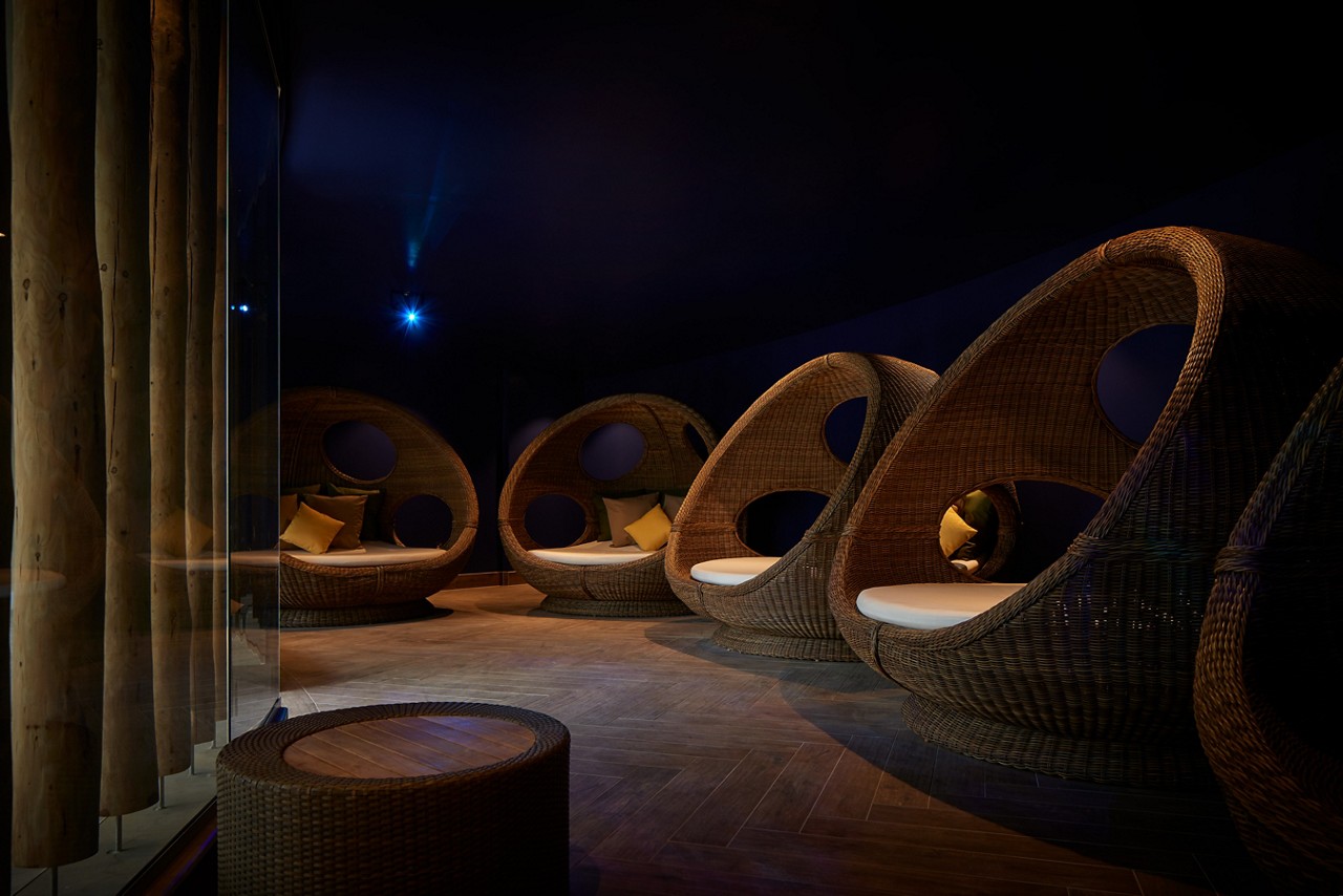 Wicker pod chairs cradle cushions, arranged in a dim lounge; soft spotlights glow against dark walls, with wood flooring and log-like columns, creating a quiet relaxation space.
