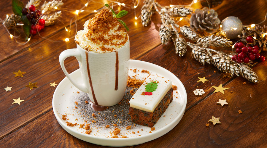 A festive hot chocolate topped with whipped cream and gingerbread crumbs served with a slice of cake.