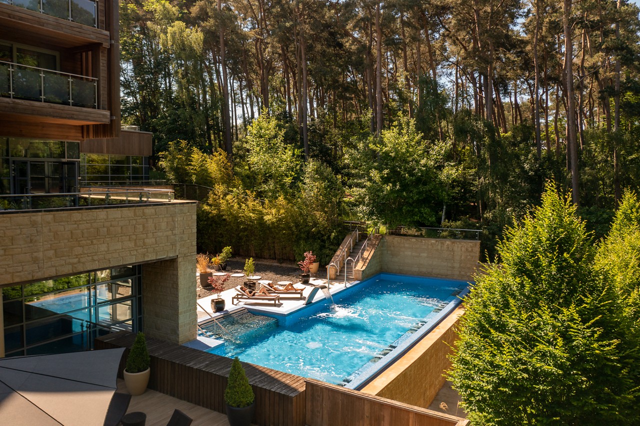 Rectangular outdoor pool bubbles with jets, steps descending into bright blue water; sun loungers and patio sit nearby, adjoining a modern stone-and-glass building, surrounded by tall pine trees.