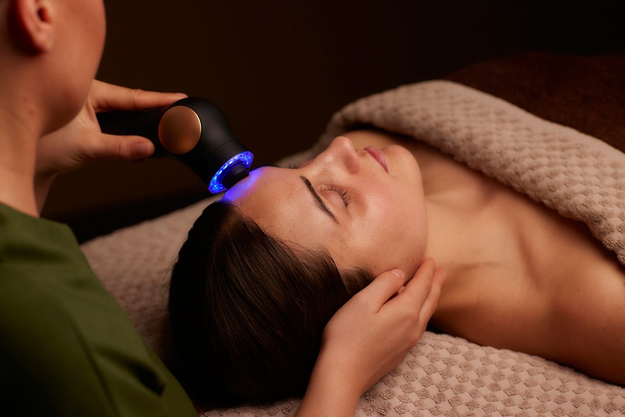 LED device shines blue light on a person's forehead as another hand supports their head. They lie on a spa bed under a textured blanket in a dim, relaxing room.