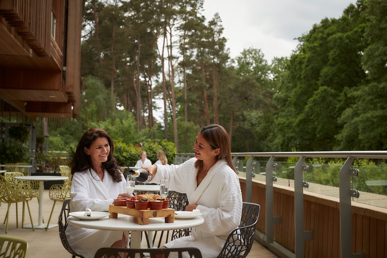 Two people in white robes pour sparkling drink at a small outdoor table, sharing appetizers. Surrounded by forested trees, metal railings, and patio seating beside a wooden building.