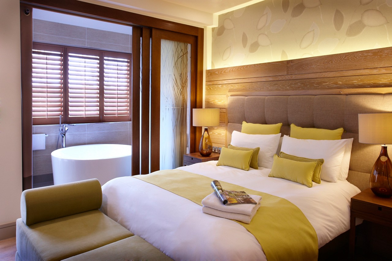 A luxury hotel bedroom with a large double bed dressed in white and yellow linens, featuring an open plan view into an ensuit bathroom with a freestanding circular bathtub