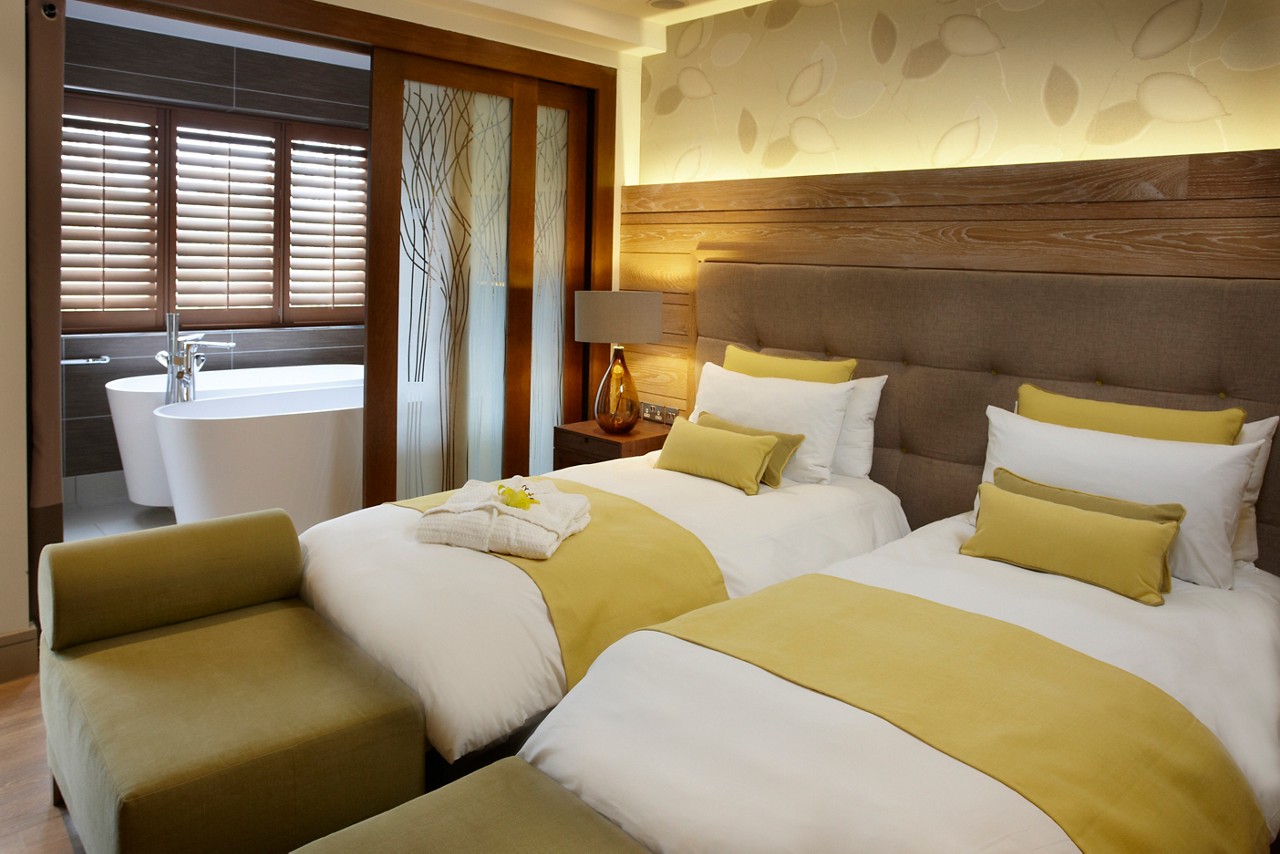 A luxury hotel bedroom with a large double bed dressed in white and yellow linens, featuring an open plan view into an ensuit bathroom with a freestanding circular bathtub