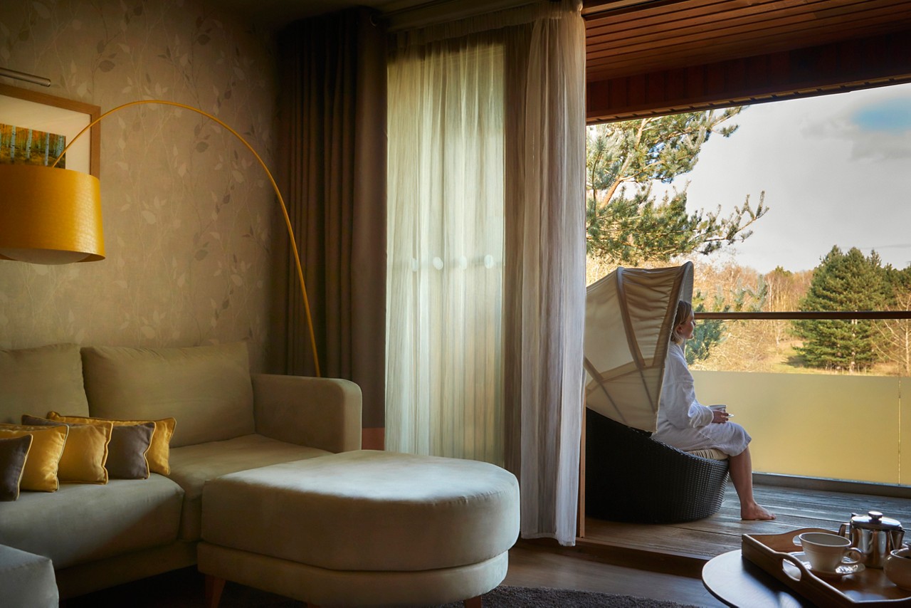 Person sits in a canopy lounge chair, sipping from a cup. They face a balcony view of trees, beside a living room with sofa, yellow arc lamp, and sheer curtains.