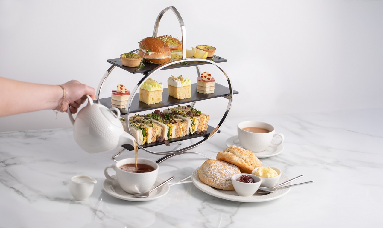 three tiered serving stand filled with sandwiches, cakes and pastries. a hand pouring tea from a white ceramic teapot into a tea cup. a plate holds scones served with jam and clotted cream
