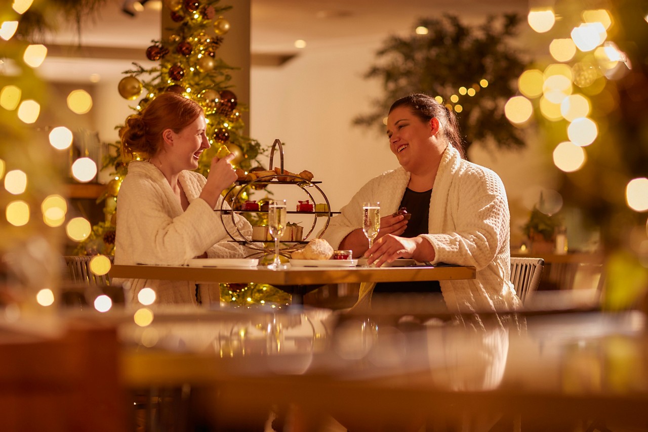 two women in spa robes sat at a wooden table with christmas decorations and lights around them
