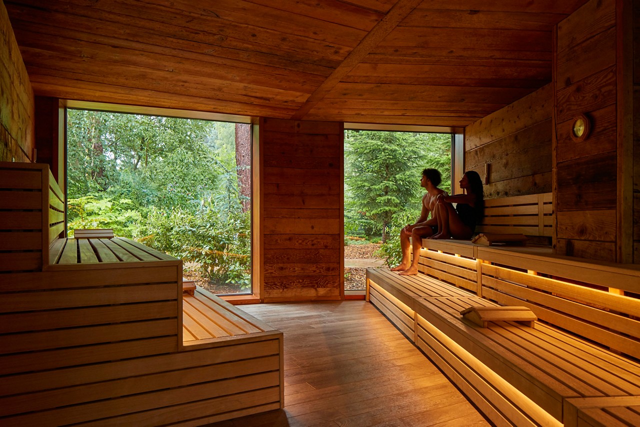 Two people sit relaxing on tiered wooden benches, gazing through large windows. Warm under-seat lighting glows. Context: a spacious cedar sauna overlooking lush forest trees and garden outside.