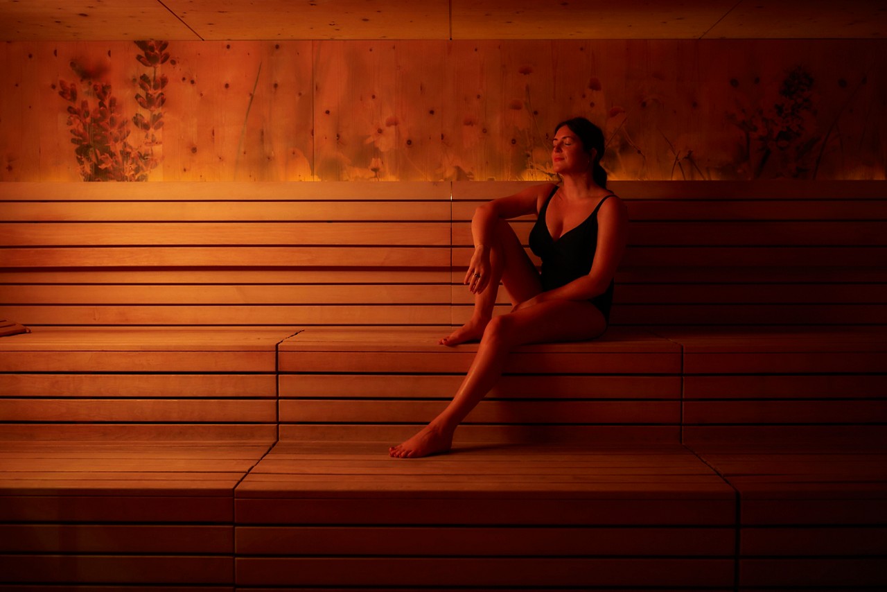 Person sits relaxed in a swimsuit, resting on tiered wooden benches, eyes closed. Warm orange lighting illuminates a sauna interior with floral silhouettes on wood paneling.