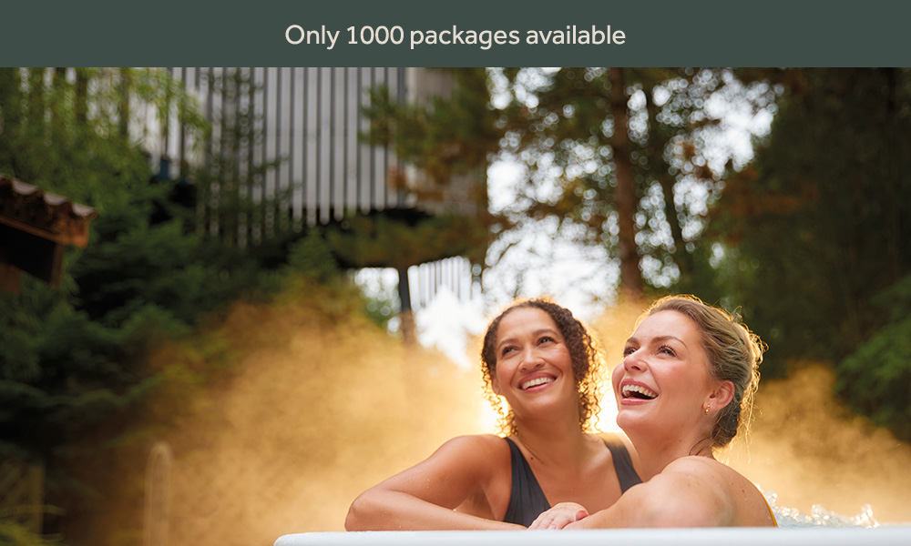 two women in an outdoor hot tub smiling with forest trees behind them