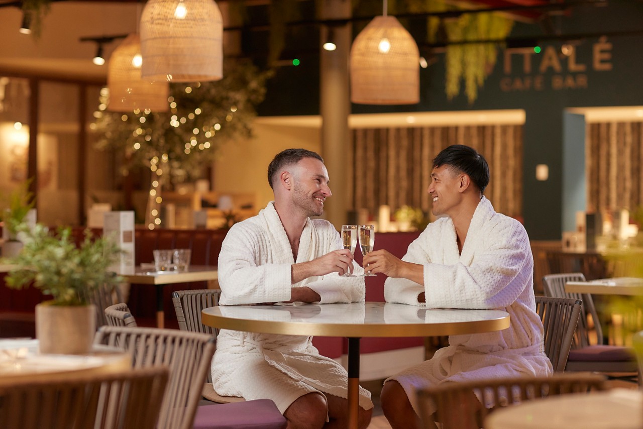 two men in spa robes sat at a table with glasses of prosecco