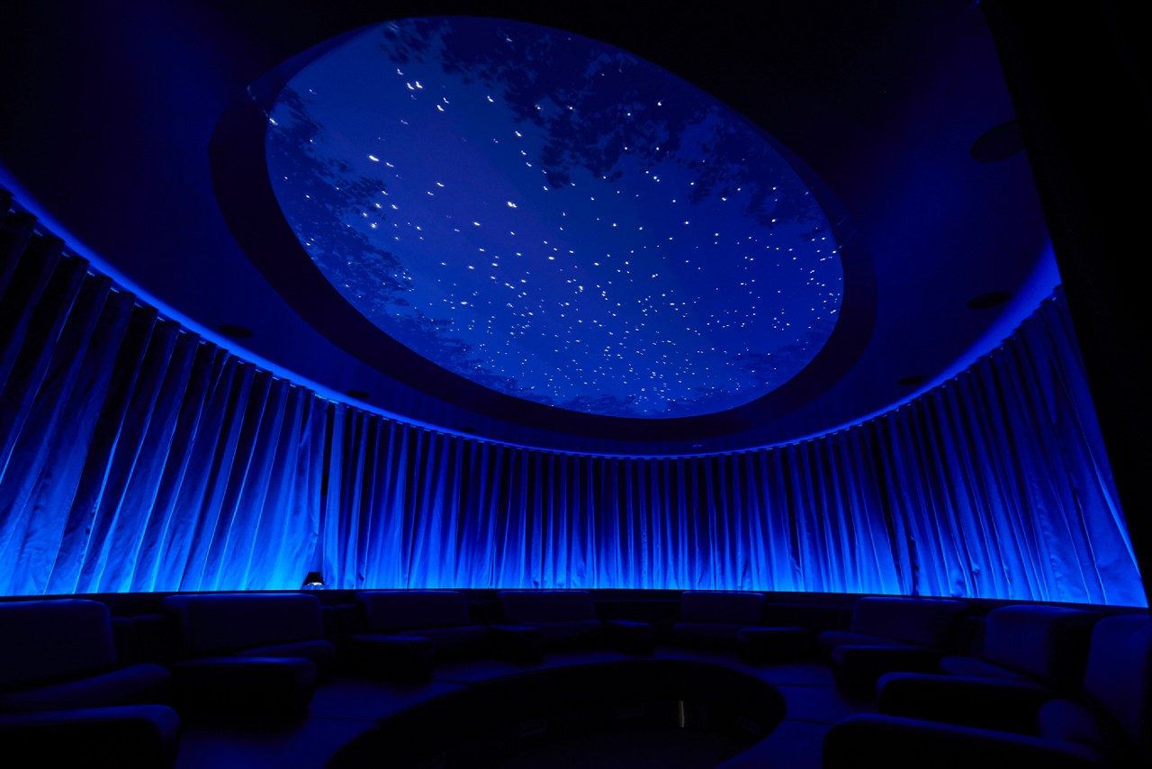 Starry oval ceiling glows, projecting constellations; circular lounge seating sits beneath; blue-lit draped walls surround in a dim, immersive room.