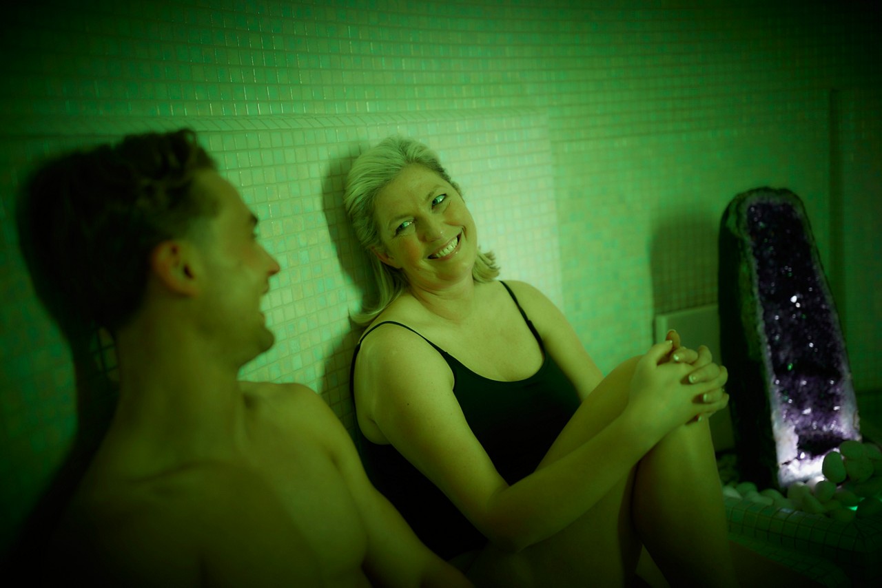 Two adults sit relaxing, smiling and chatting, in swimsuits. They lean against tiled walls under green light; a lit amethyst geode water feature stands beside them among white pebbles.