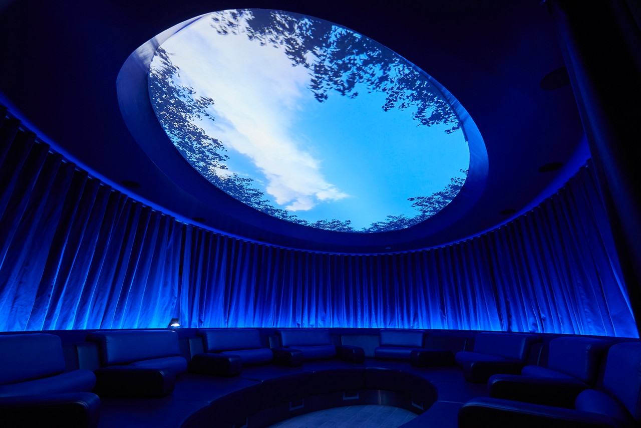 Circular lounge seating encircles a sunken floor, illuminated blue; above, an oval skylight reveals sky and tree silhouettes, within a dark, draped interior.