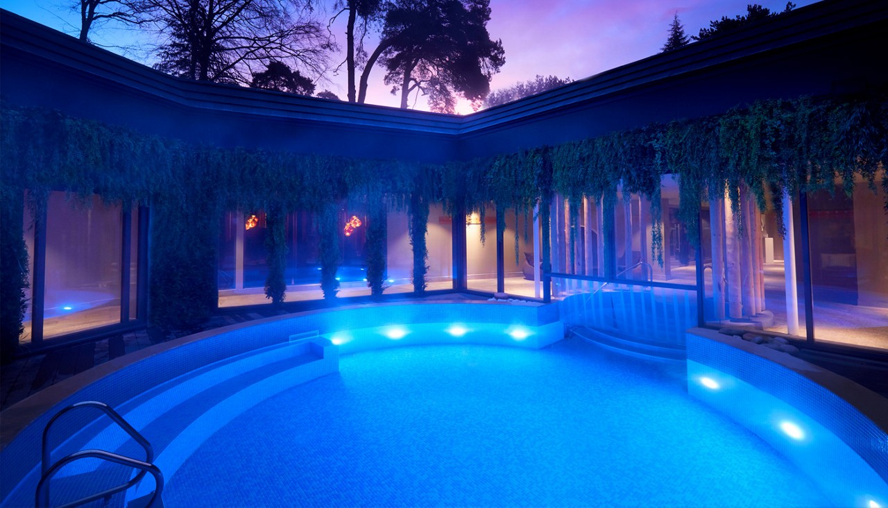 Curved swimming pool glowing blue, illuminated underwater lights, unused at dusk; indoor-outdoor spa courtyard with glass walls, hanging plants, steps and metal handrail; trees silhouetted against purple sky.