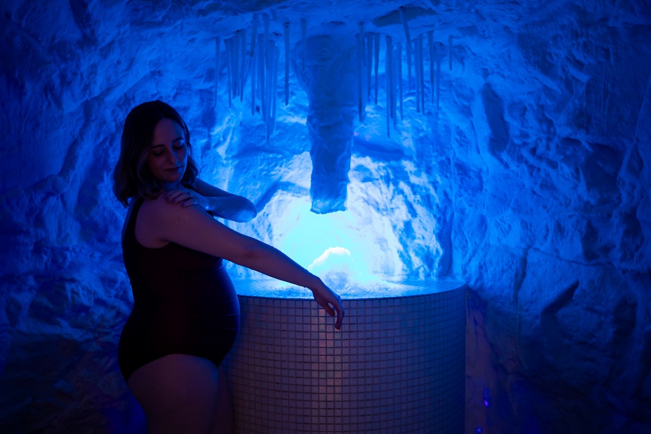 Person in a dark swimsuit rests an arm on a rounded tiled hot tub, lightly touching their shoulder, in a blue-lit cave-like room with icicles and a glowing water feature.