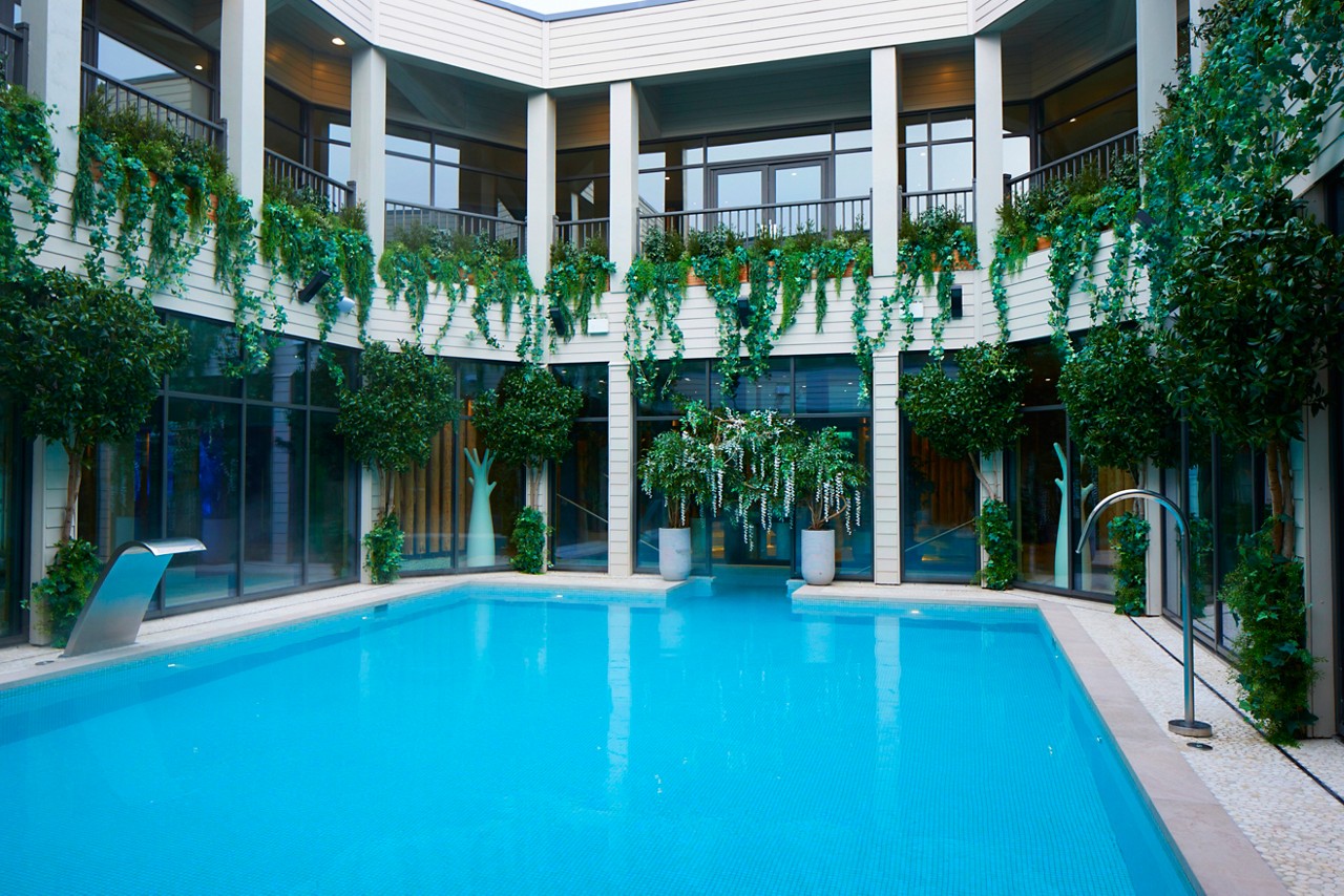 Swimming pool gleams calmly, reflecting light; metal fountains flank edges. Context: modern two-story courtyard with glass walls, balconies, and abundant hanging vines and potted trees surrounding the serene aqua water.