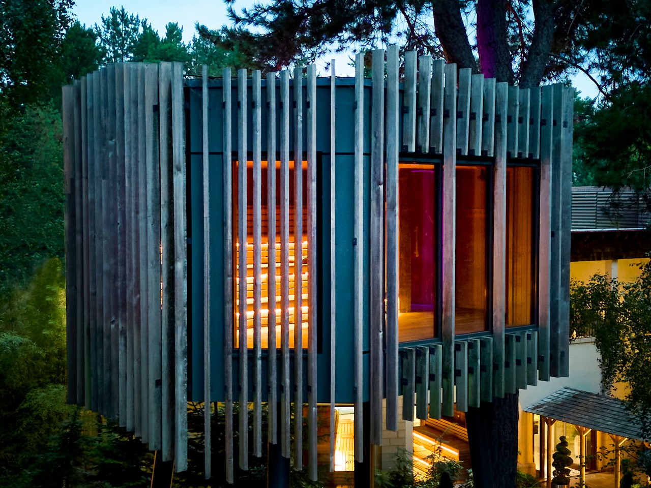 Modern treehouse glows with warm interior light, perched on tall stilts; large windows reveal wooden benches. Dense forest at dusk surrounds it, with nearby illuminated buildings and purple accent lighting.