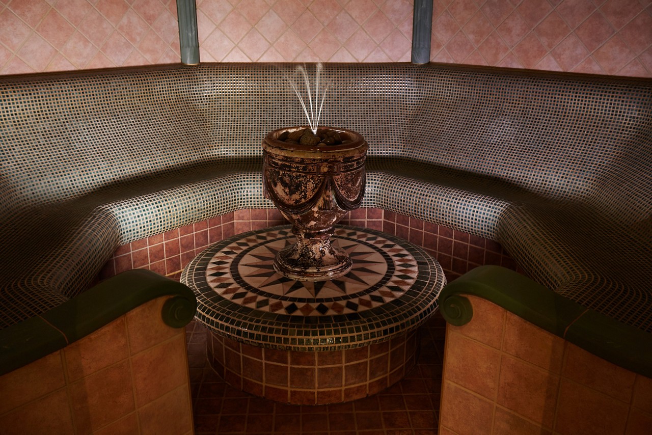 Ornate urn fountain sprays thin water jets upward, resting on a mosaic pedestal. Surrounding it, a curved, tile-covered bench forms a circular steam-room alcove with warm, dim lighting.
