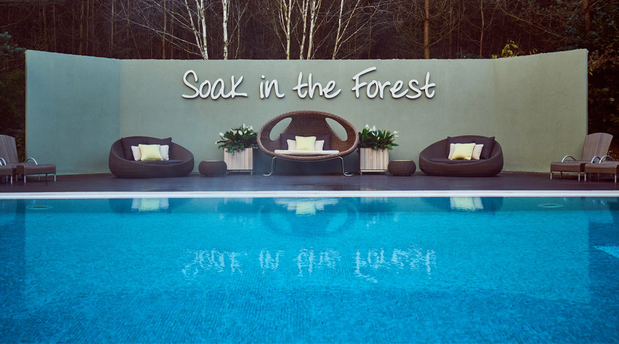 Outdoor pool with surrounding decking where comfortable egg chairs are placed in front of a sign that reads 'Soak in the Forest'.