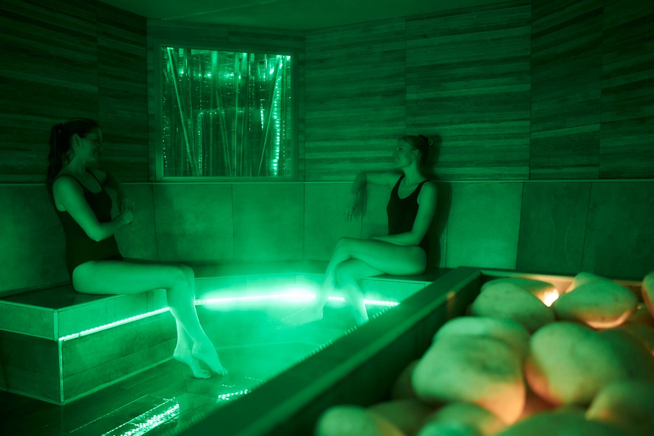 Two people in swimsuits sit and relax on tiled benches, feet near a shallow illuminated pool, in a green-lit steam room with hot stones glowing in the foreground.