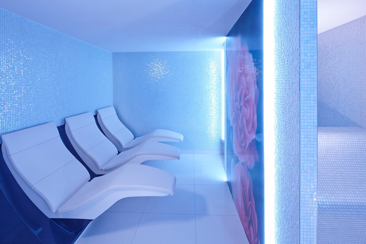 Four white reclining lounge chairs sit empty; in a blue-lit spa room with mosaic tile walls and floor; a translucent panel with rose artwork separates an adjacent tiled alcove.
