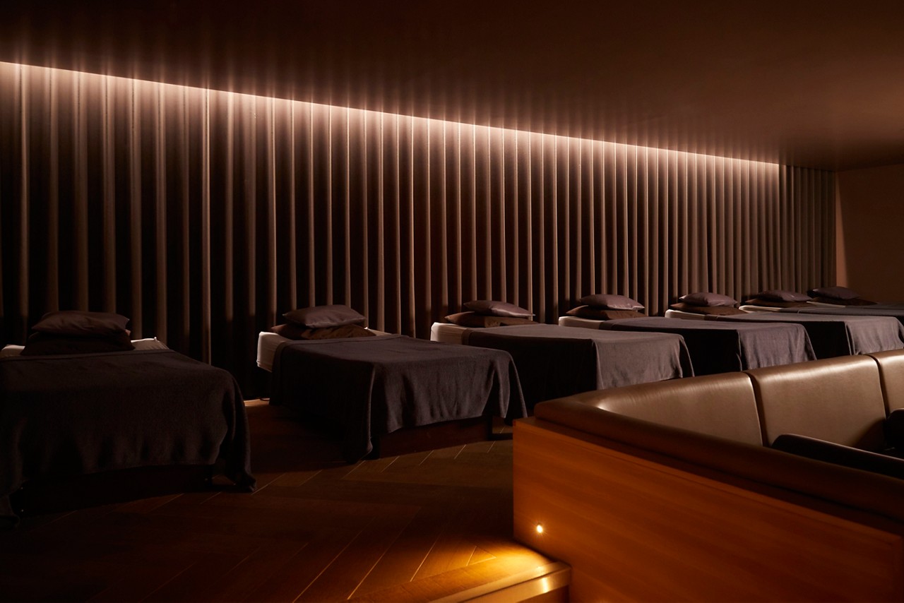 people in spa robes lying on padded daybeds resting. Warm, low lighting and ribbed wall panels create a calm, dim relaxation-room ambiance.