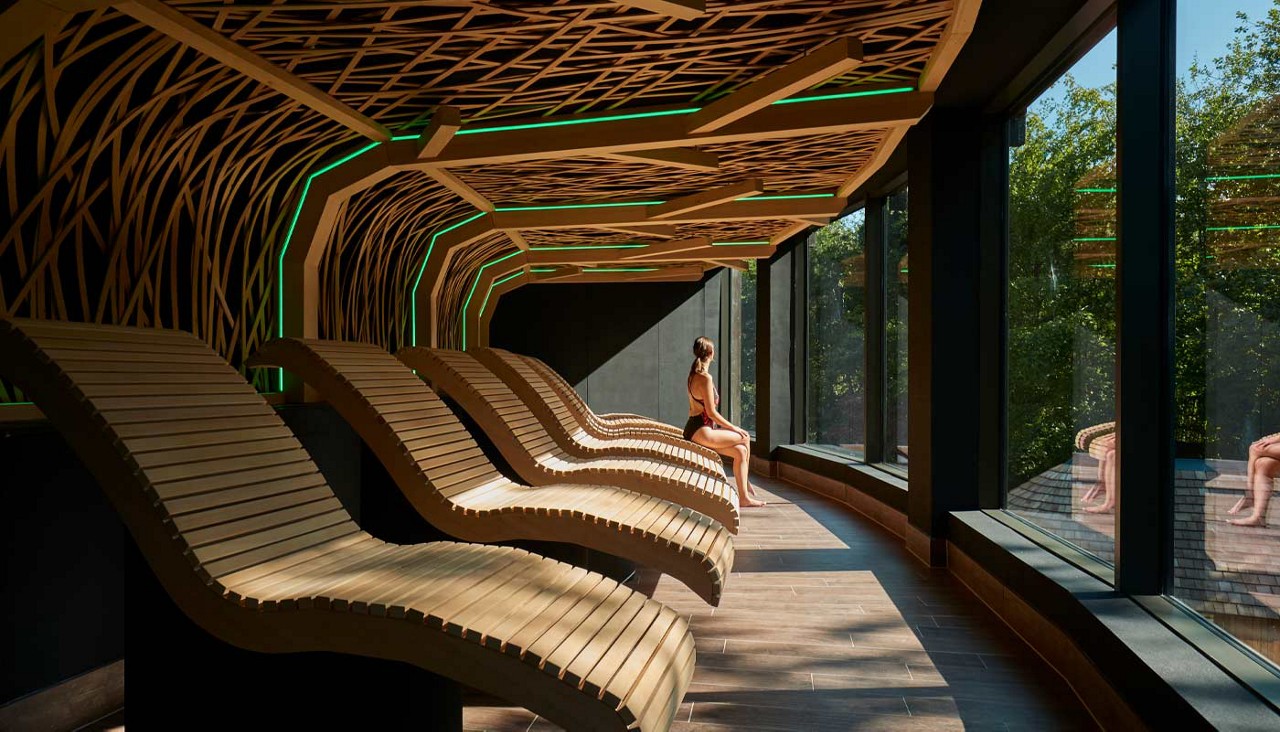 Curved wooden loungers line a room while a seated person relaxes, facing floor-to-ceiling windows; organic lattice ceiling with green accent lights frames a serene forest view outside.