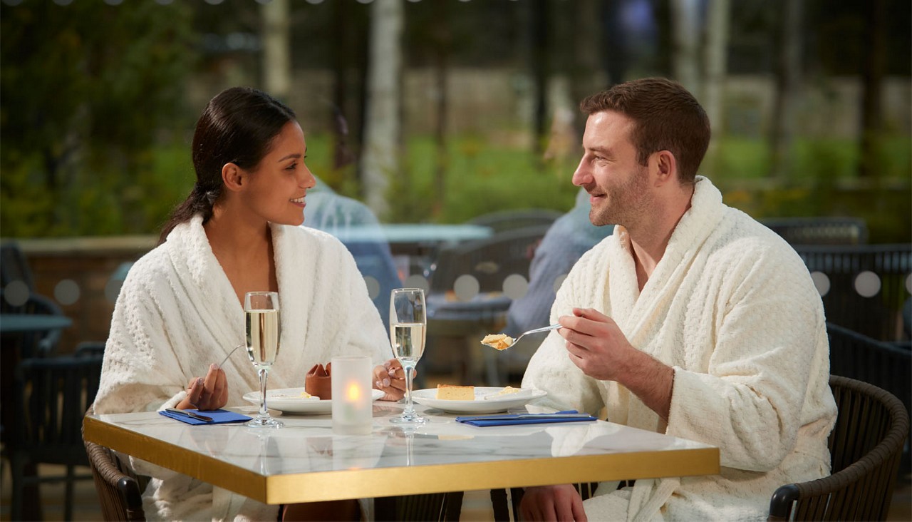 Two adults in white bathrobes converse and eat dessert, one lifting a spoon, at a small outdoor cafe table with champagne flutes, plates, and a candle, greenery blurred in background.