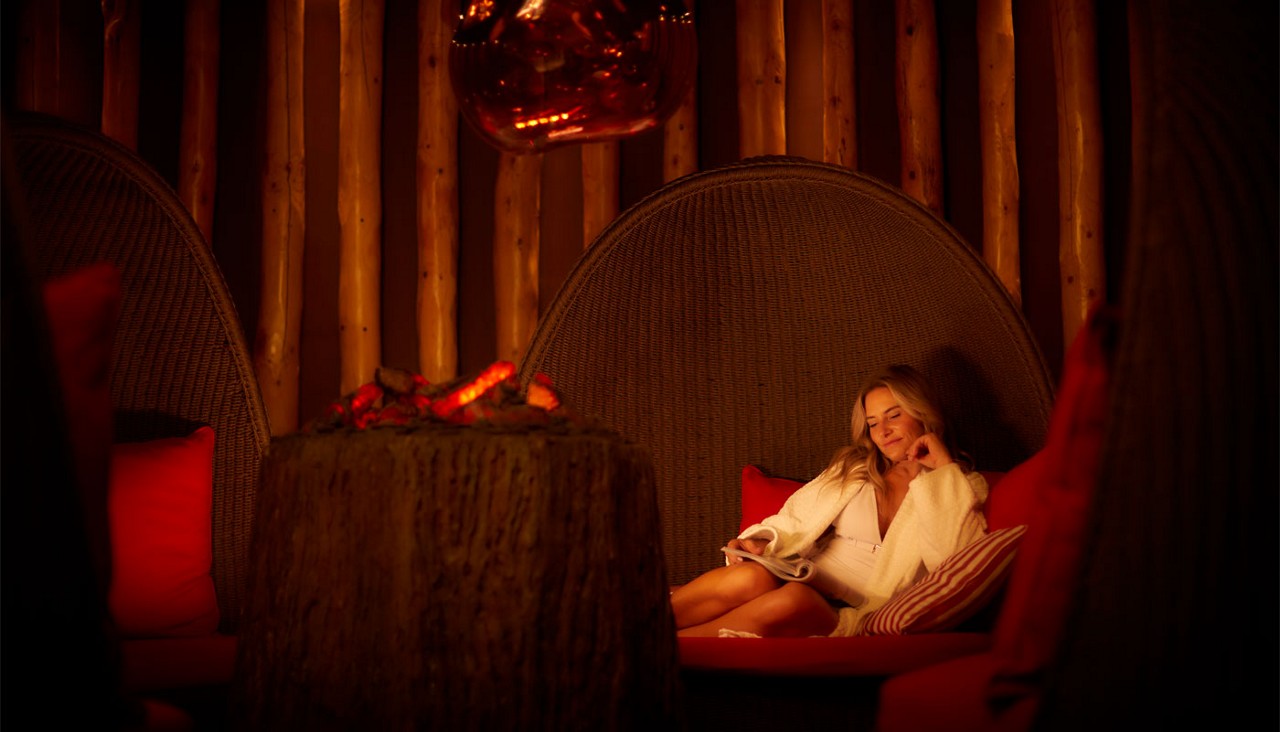 Woman reclines and smiles in a dome-shaped wicker lounge, holding a book, as a glowing fire pit flickers nearby within a warmly lit, wooden, cabin-like room.