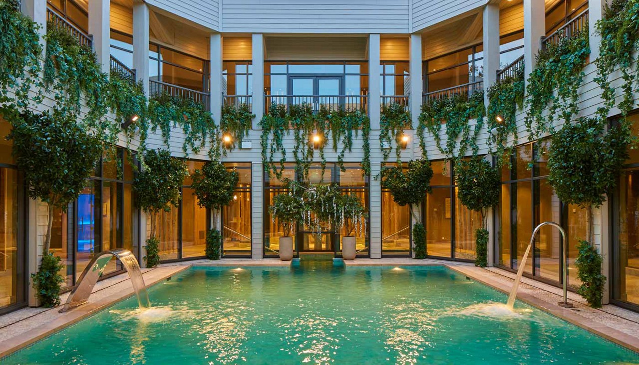 Pool with jets spouting into turquoise water, centered in a courtyard enclosed by a two-story building with glass walls, balconies, warm lights, and abundant hanging plants and potted trees.