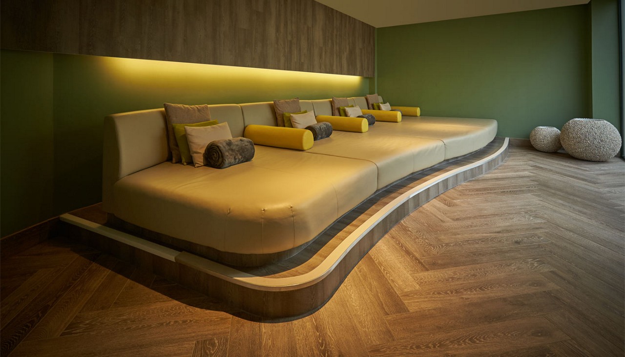 Curved, tiered lounge platform holds beige cushions with yellow bolsters and gray rolls, softly lit by wall LED strip; in a room with green walls, herringbone wood floor, pebble stools.
