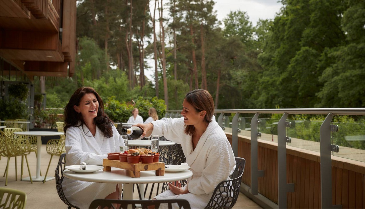 Two people in white bathrobes share brunch, one pouring sparkling wine into flutes, seated at a round patio table with pastries, on a balcony overlooking lush trees and forest.