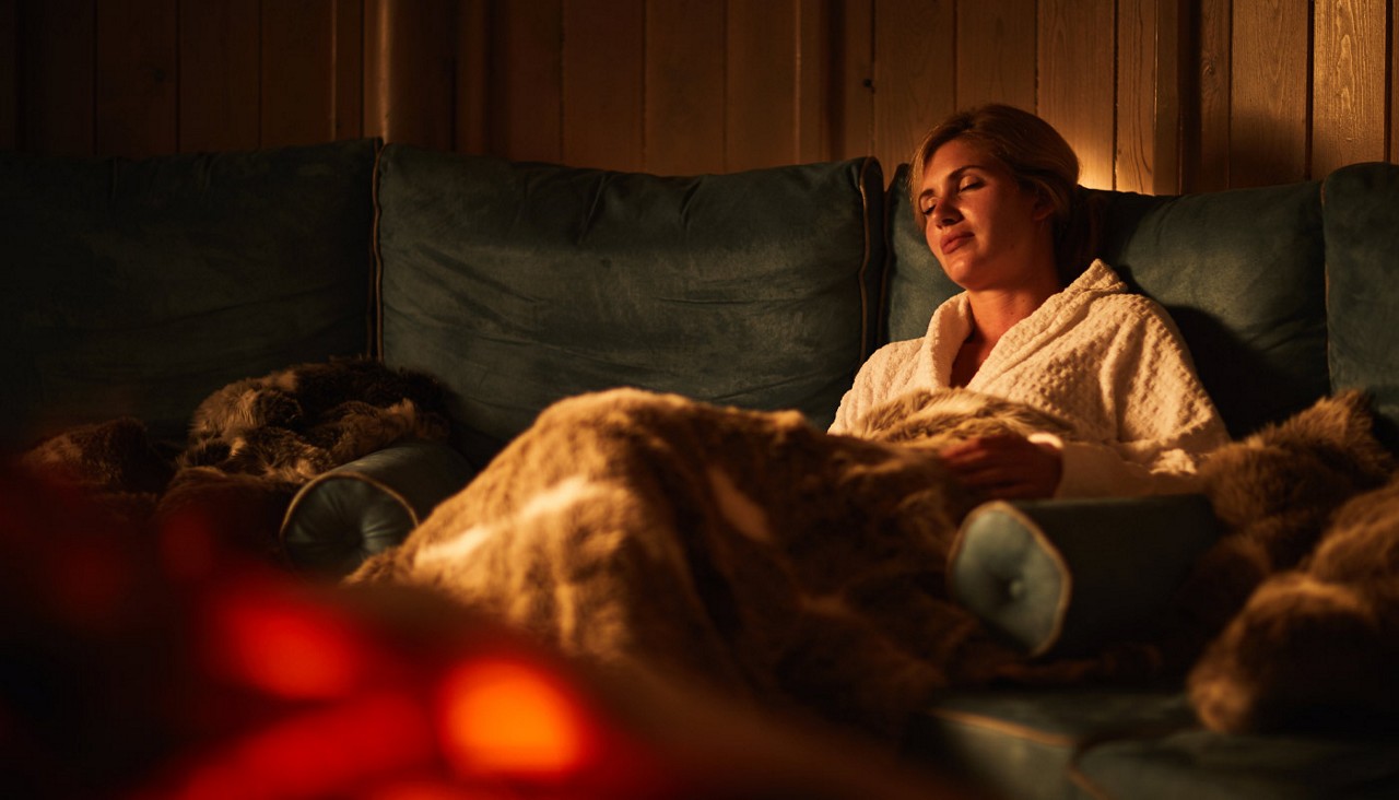 Woman snuggled under a cosy blanket beside a glowing fire.