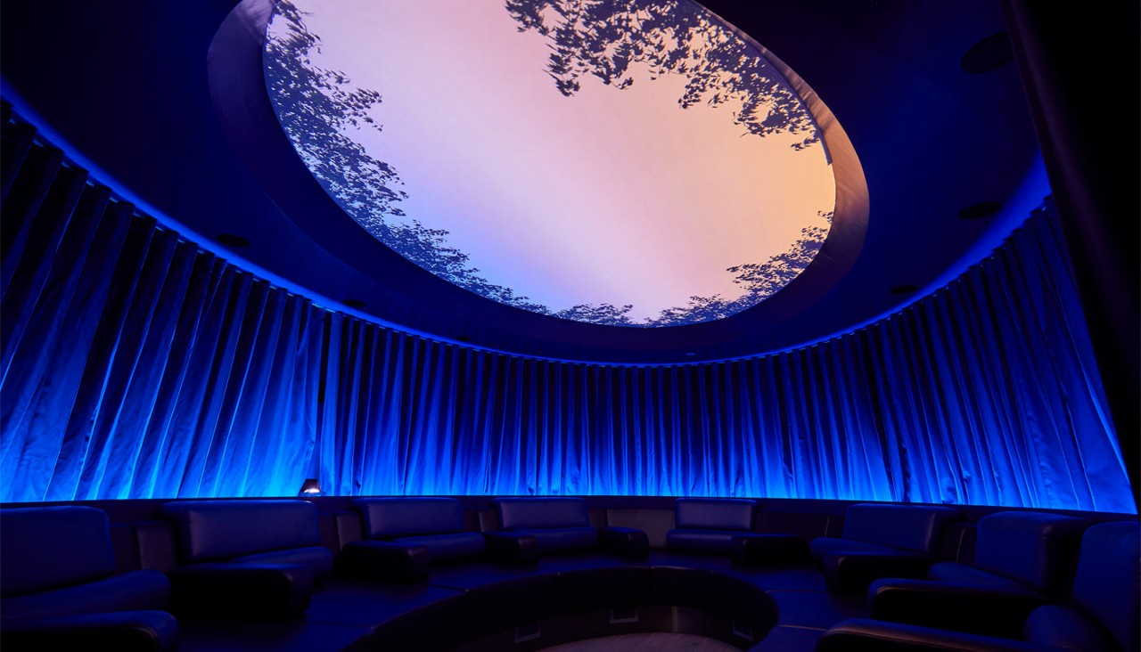 Skylight displays pastel sky and tree silhouettes, casting light while encircling sofas sit below in a dim, circular room with pleated blue-lit curtains and a central sunken floor.