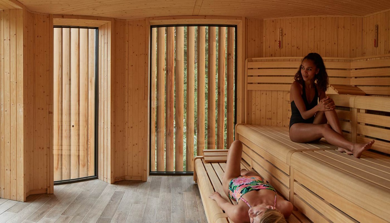 Two people relax—one sitting, one lying—on tiered wooden benches inside a modern sauna. Tall vertical-slat windows admit soft light, and warm wood walls and floor surround them.