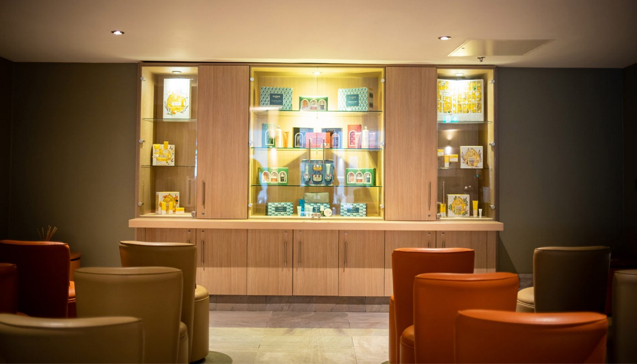 Illuminated wooden display cabinet showcases colorful boxed gift sets on glass shelves, while rounded leather chairs sit arranged in front, suggesting a quiet lounge or boutique waiting area.
