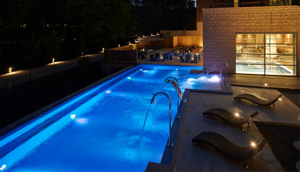 Illuminated swimming pool glows blue, bubbles, and sprays from stainless arches; steps descend. Nearby curved loungers line a patio as an adjacent glass-walled building reveals an indoor pool at night.