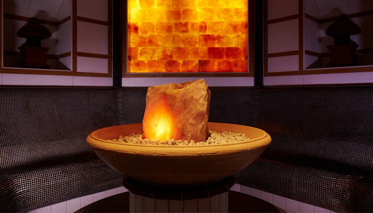 Glowing salt rock lamp lights a shallow pebble-filled bowl on a pedestal. Curved benches with dark mosaic tiles surround it in a sauna or steam room, backed by amber-lit wall.
