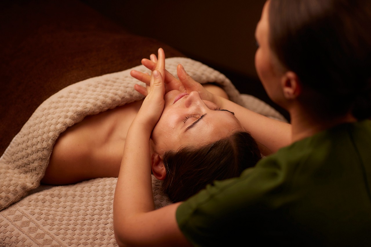 A spa therapist giving a facial treatment to client lying on a spa bed