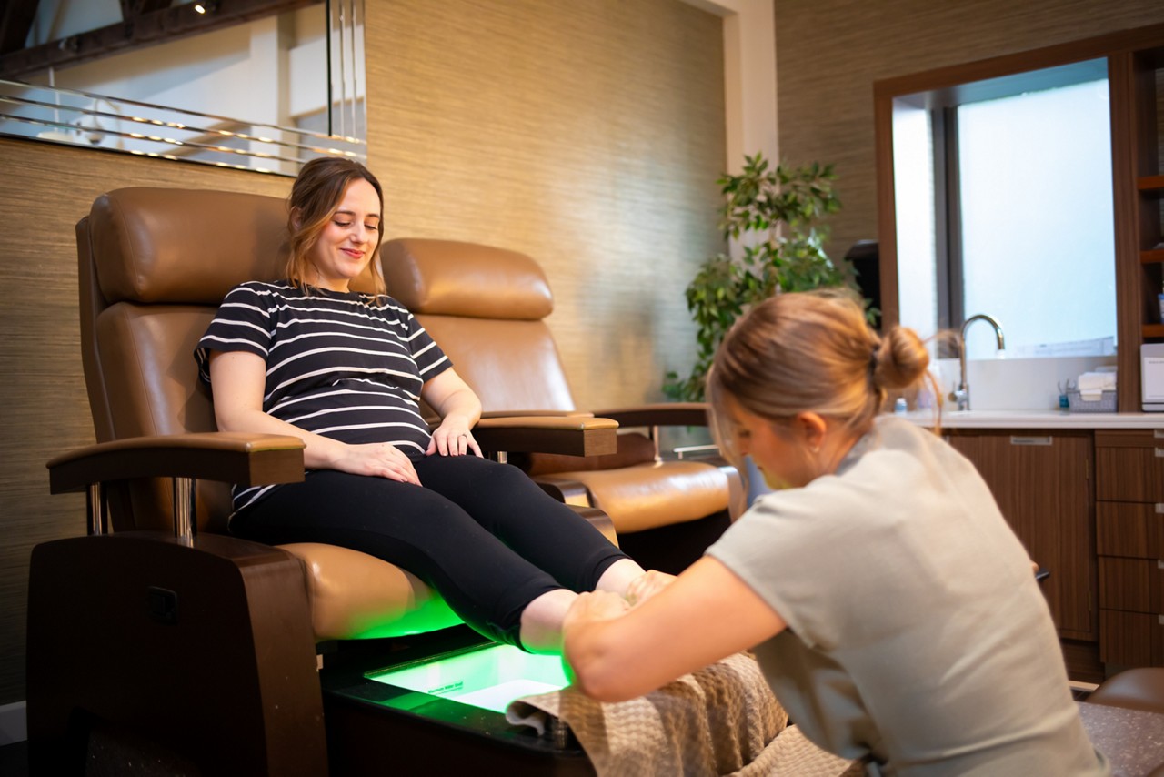 Client reclines on a pedicure chair, receiving a foot massage from a technician, in a modern spa with brown leather seats, glowing green footbath, shelves, sink, and vanity lights.