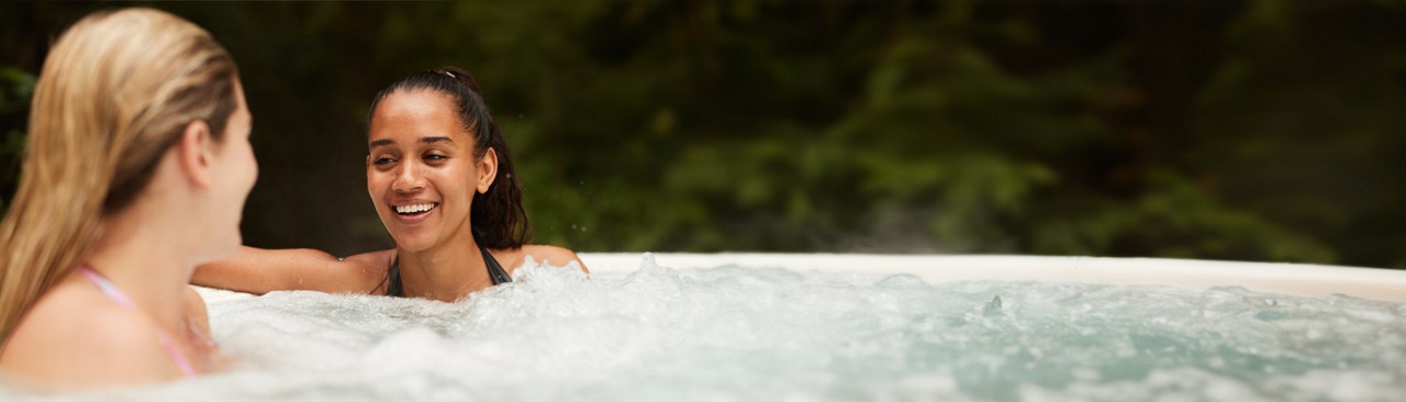 Two women sitting in a bubbling hot tub surrounded by the forest.